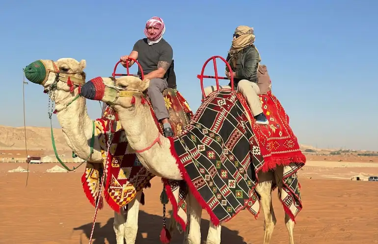Two tourists wearing traditional headscarves riding camels decorated with vibrant traditional saddles during a desert tour at Red Sand Desert in Riyadh.