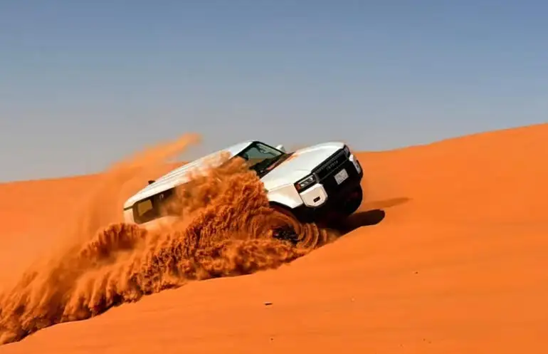 A white Toyota SUV performing dune bashing and kicking up sand on a steep slope in the Red Sand Desert of Riyadh.