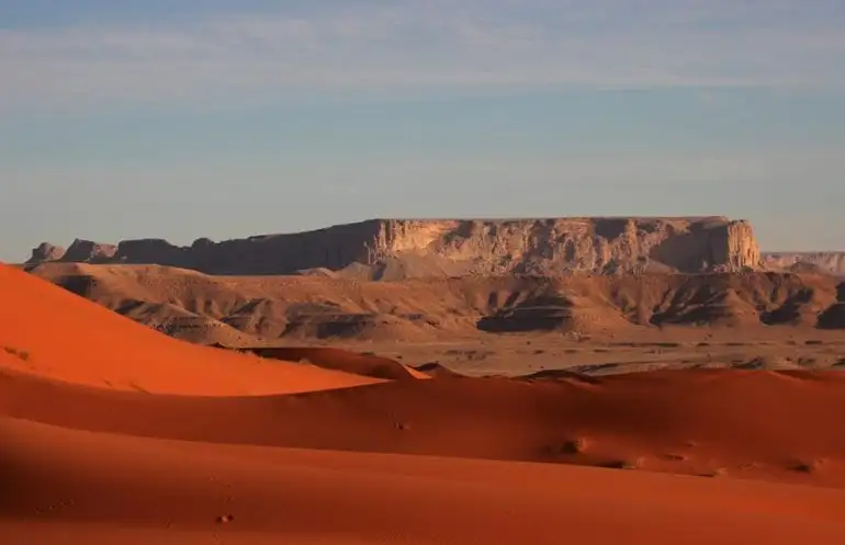 A wide panoramic view of rolling red sand dunes in the foreground with rugged sandstone cliffs and plateaus in the distance under a clear sky in Riyadh.