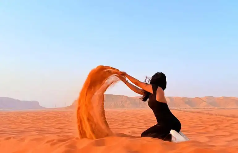 A woman kneeling on a vibrant orange dune and throwing a handful of sand into the air at the Red Sand Desert near Riyadh, Saudi Arabia.