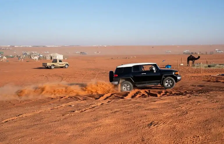 Black 4x4 off-roading in Riyadh Red Sand Desert with camels and desert camp in background