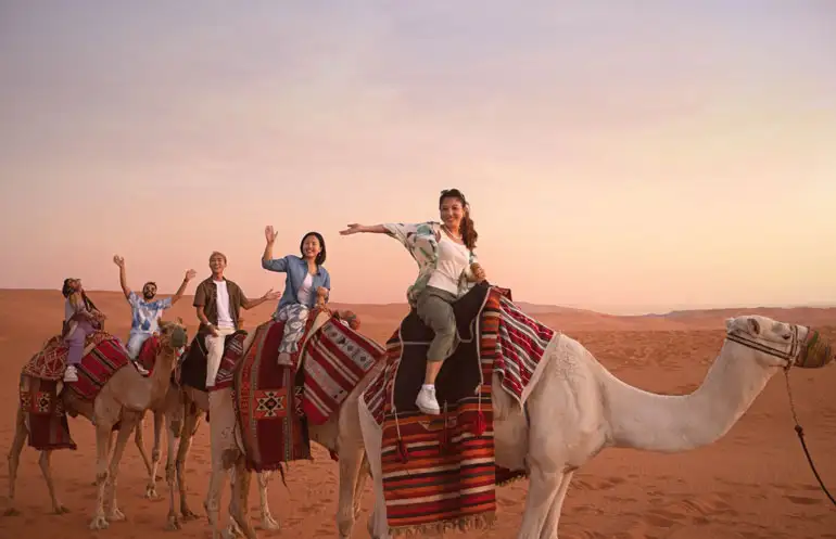 Happy group of friends and family riding camels through the stunning red sand dunes at golden hour during the Red Sand Tour in Riyadh. A memorable desert experience for all ages.