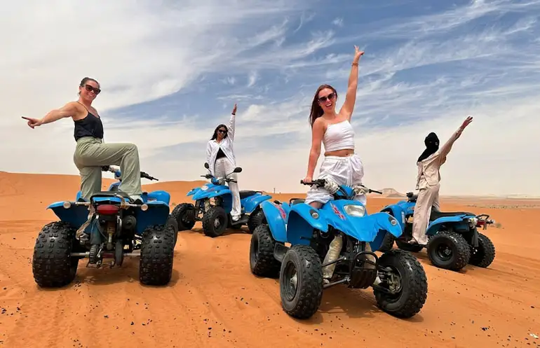 A group of four women smiling and posing energetically on blue all-terrain vehicles (ATVs) in the vibrant red sand dunes of Riyadh's Red Sand Desert. One woman in a black top points forward, another in white raises her arm, and the others wave joyfully under a bright blue sky.