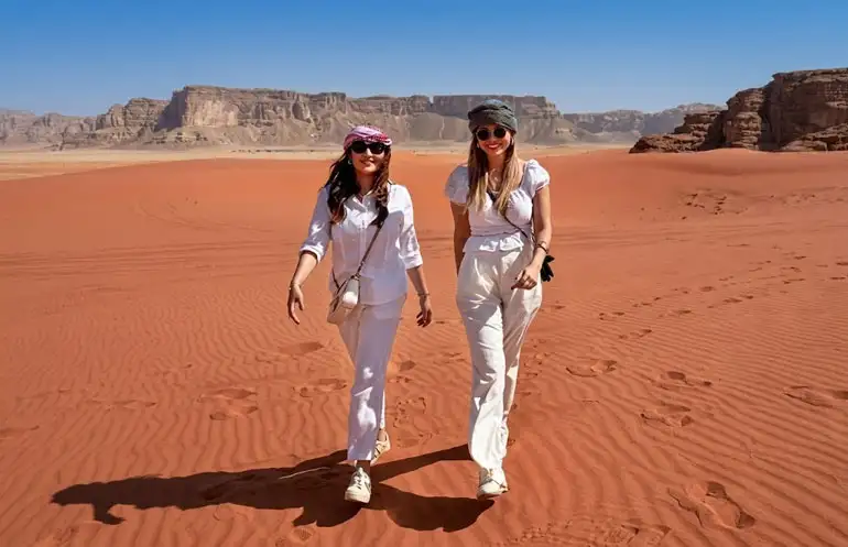 Two women dressed in stylish white outfits and sunglasses walking side-by-side across the rippled red sand dunes of Riyadh's Red Sand Desert, with dramatic rocky cliffs visible in the background under a clear blue sky.