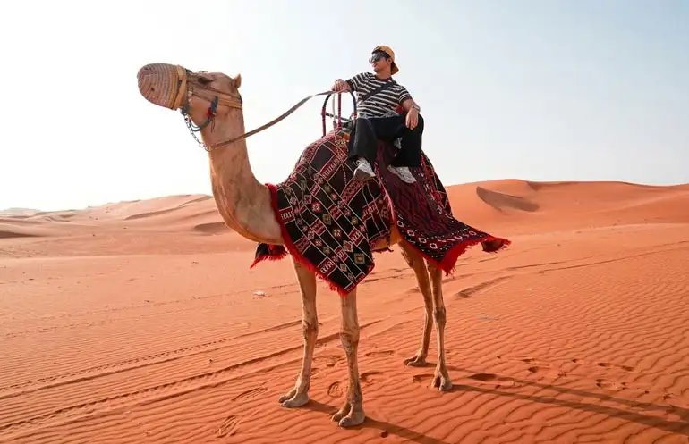 A man wearing a striped shirt and cap riding a decorated camel across the orange-red sand dunes of Riyadh's Red Sand Desert, with vast sandy landscape and clear sky in the background.